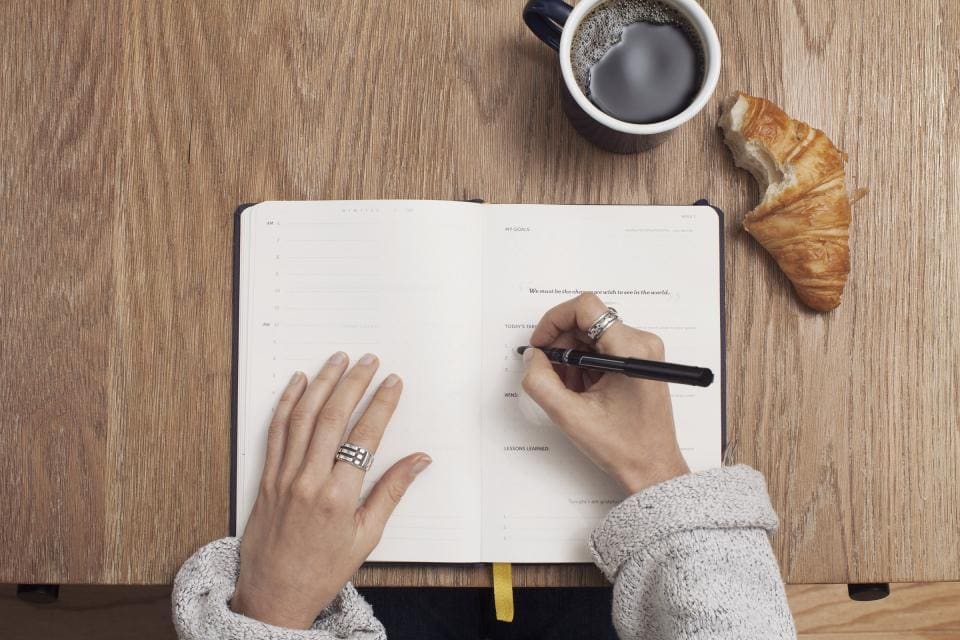 Hands writing in an open notebook on a wooden table, with a cup of coffee alongside